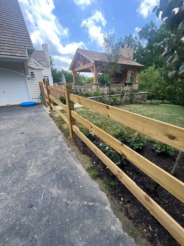 Three-board pressure-treated post and rail fence running along a driveway toward a stone pavilion in Wayne, PA